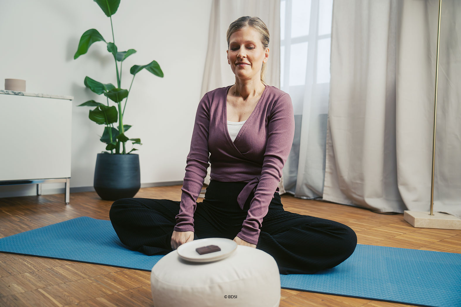 Eine Frau sitzt in Yoga-Position vor einem textilen Rundhocker, auf dem ein Tellerchen mit Schokolade platziert ist. Offenbar freut sich die Frau auf den bevorstehenden Schokoladengenuss.